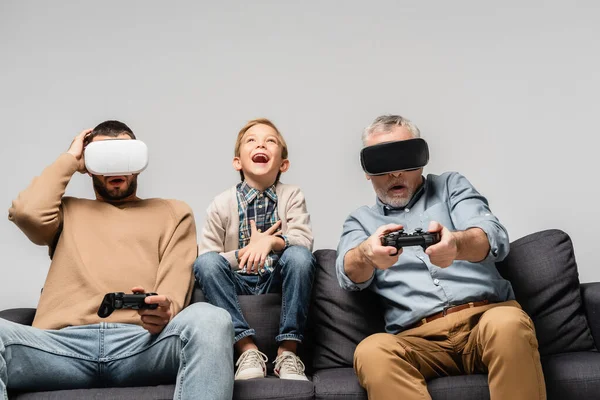 KYIV, UCRANIA - 17 de noviembre de 2020: niño emocionado riéndose cerca de papá y abuelo jugando videojuegos en auriculares vr aislados en gris - foto de stock