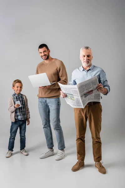 Homme mature tenant journal près de fils et petit-fils avec des gadgets sur gris — Photo de stock