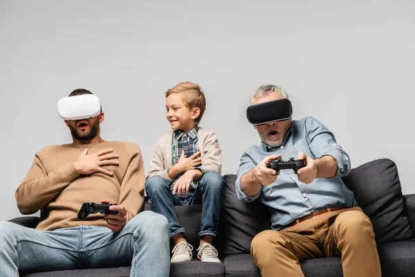 KYIV, UKRAINE - NOVEMBER 17, 2020: cheerful boy near scared dad and grandfather playing video game in vr headsets isolated on grey — Stock Photo
