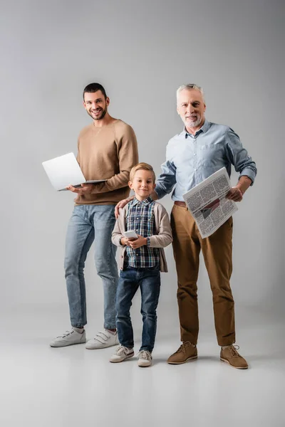 Niño alegre sosteniendo teléfono inteligente cerca del abuelo con el periódico y papá con el ordenador portátil en gris - foto de stock