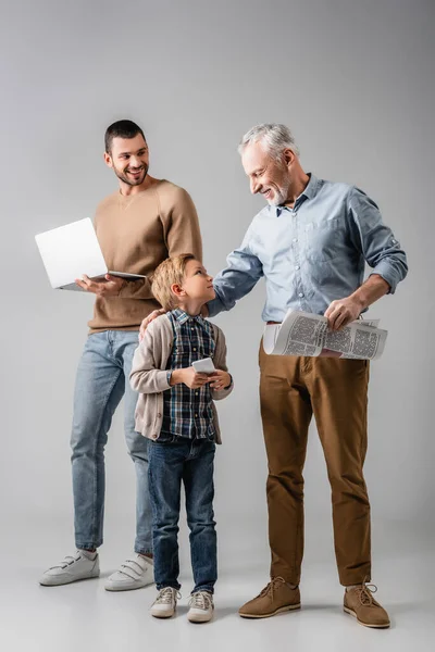 Niño feliz mirando al abuelo con el periódico cerca de papá sosteniendo portátil en gris - foto de stock