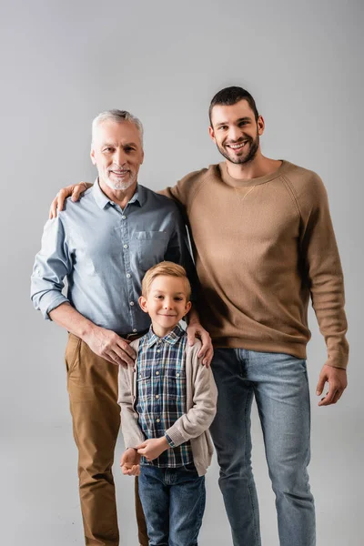 Щасливий хлопчик дивиться на камеру біля батька і дідуся ізольовано на сірому — стокове фото