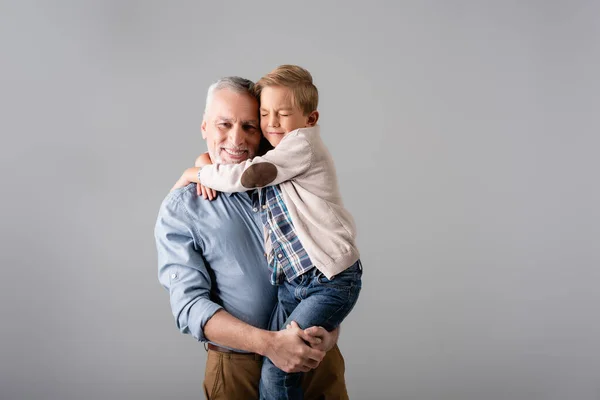 Alegre hombre mirando a la cámara mientras sostiene nieto abrazándolo con los ojos cerrados aislados en gris - foto de stock