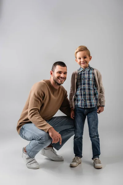 Happy father squatting near cheerful son while smiling at camera together on grey — Stock Photo