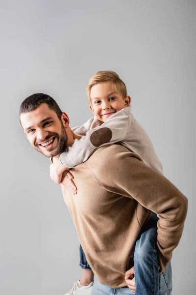 Felice uomo piggybacking sorridente figlio mentre guardando la fotocamera insieme isolato su grigio — Foto stock