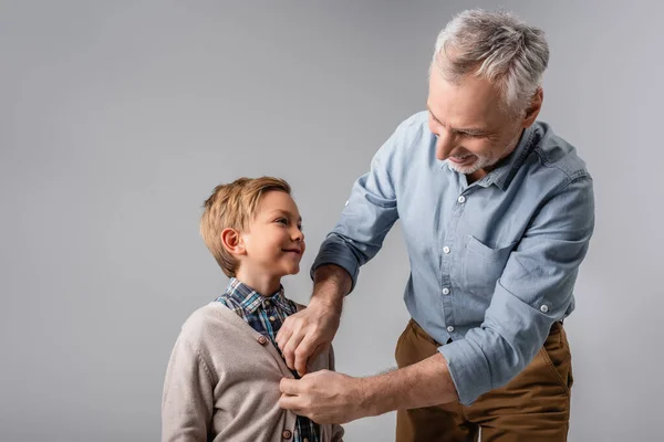 Дедушка застегивает рубашку улыбающегося внука, изолированного на сером — стоковое фото