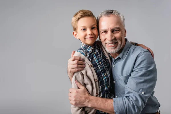 Happy mature man embracing smiling grandson while looking at camera isolated on grey — Stock Photo