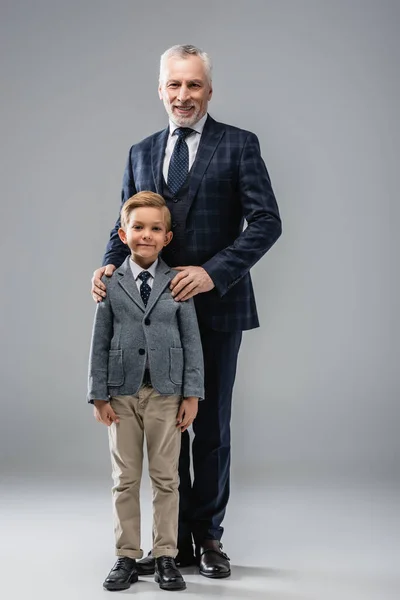 Happy businessman hugging shoulders of grandson while standing on grey — Stock Photo