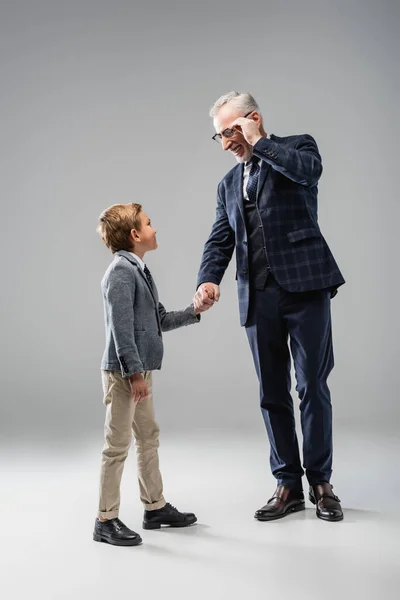 Happy businessman touching eyeglasses while holding hands with grandson on grey — Stock Photo