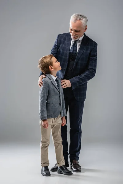 Smiling businessman in formal wear hugging shoulders of grandson on grey — Stock Photo