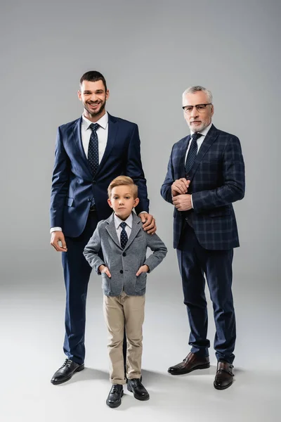 Hombres de negocios sonrientes en ropa formal mirando a la cámara cerca de niño de pie con las manos en los bolsillos en gris — Stock Photo