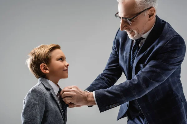 Happy businessman in eyeglasses buttoning blazer of smiling grandson isolated on grey — Stock Photo