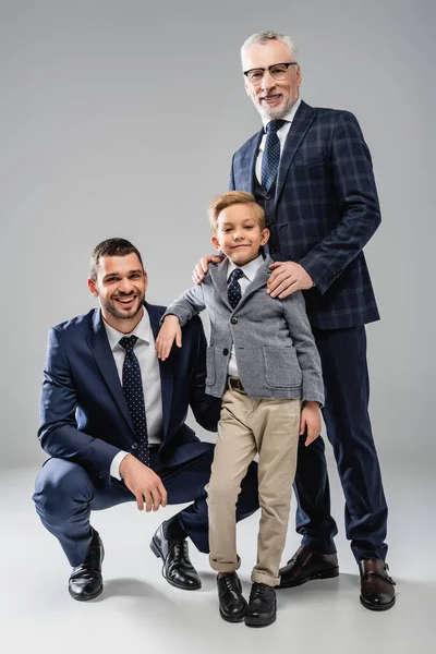 Happy businessmen with boy in formal wear smiling at camera on grey — Stock Photo