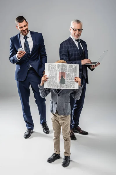 Sonrientes hombres de negocios con gadgets mirando a chico oscureciendo la cara con periódico en gris - foto de stock
