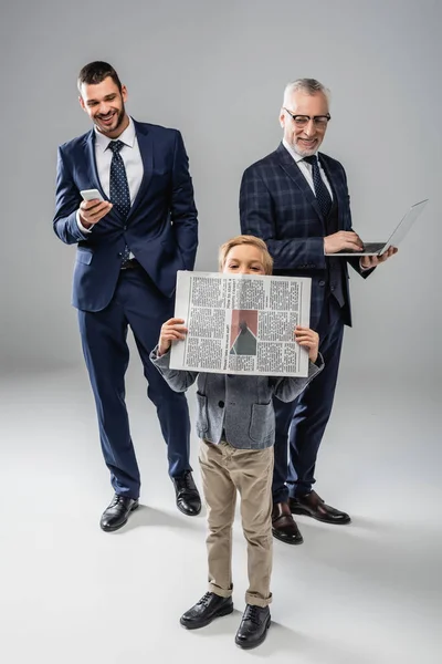 Sonrientes hombres de negocios en ropa formal sosteniendo gadgets y mirando a chico con periódico en gris - foto de stock