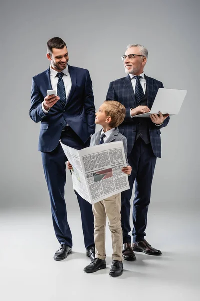 Lächelnder Junge mit Zeitung in der Nähe von Vater und Großvater, die Gadgets auf grau halten — Stockfoto