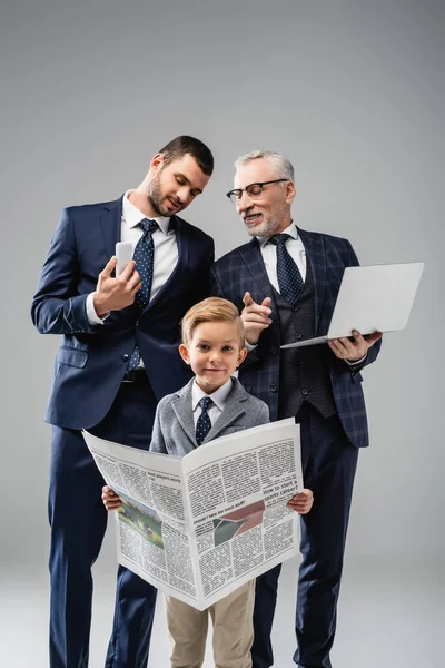 Mature businessman talking to son and pointing with finger at grandson holding newspaper on grey — Stock Photo