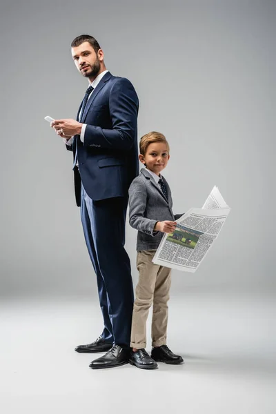 Geschäftsmann mit Smartphone und sein Sohn mit Zeitung schauen in die Kamera auf grau — Stockfoto