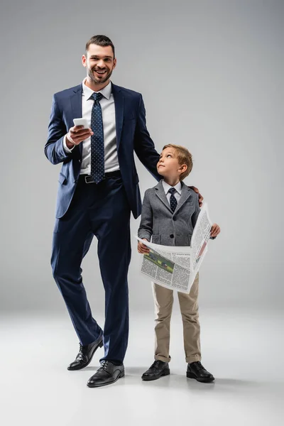 Feliz hombre de negocios con teléfono inteligente sonriendo a la cámara cerca de hijo con periódico en gris - foto de stock