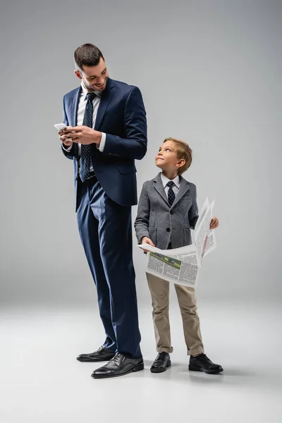 Feliz hombre de negocios con teléfono móvil mirando hijo celebración de periódico en gris - foto de stock