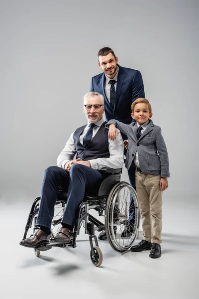 Hombre feliz en ropa formal de pie cerca de hijo y padre en silla de ruedas en gris - foto de stock