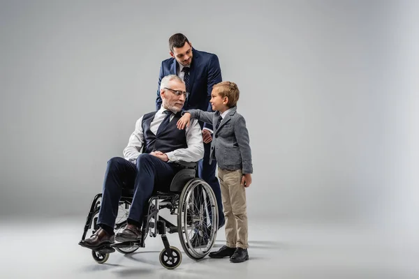 Uomo sorridente in abito formale in piedi vicino a figlio e padre in sedia a rotelle su grigio — Foto stock