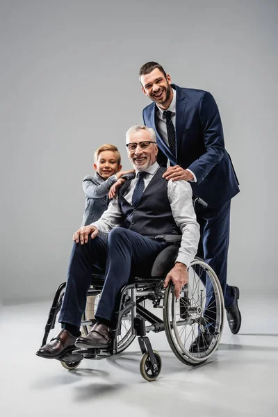 Happy man in wheelchair near son and grandson hugging his shoulders on grey — Stock Photo