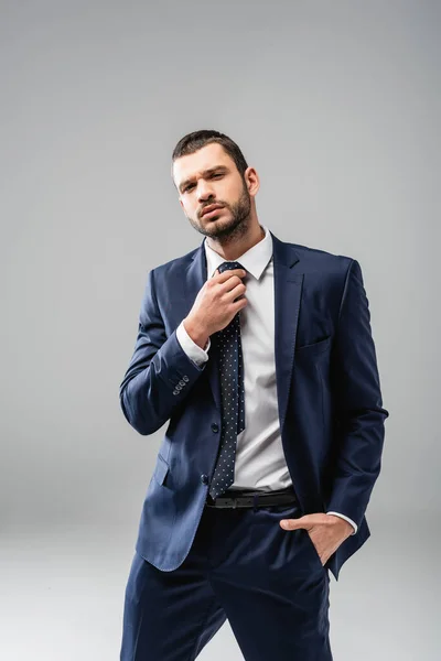 Confiado hombre de negocios fijación de corbata mientras está de pie con la mano en el bolsillo en gris - foto de stock