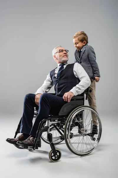 Mature man in wheelchair looking at smiling grandson on grey — Stock Photo