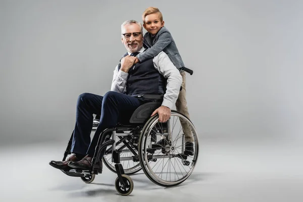 Sorridente ragazzo che abbraccia il nonno in sedia a rotelle mentre guarda la fotocamera insieme sul grigio — Foto stock
