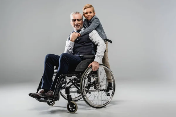 Niño sonriente abrazando al abuelo en silla de ruedas mientras miran a la cámara juntos en gris - foto de stock