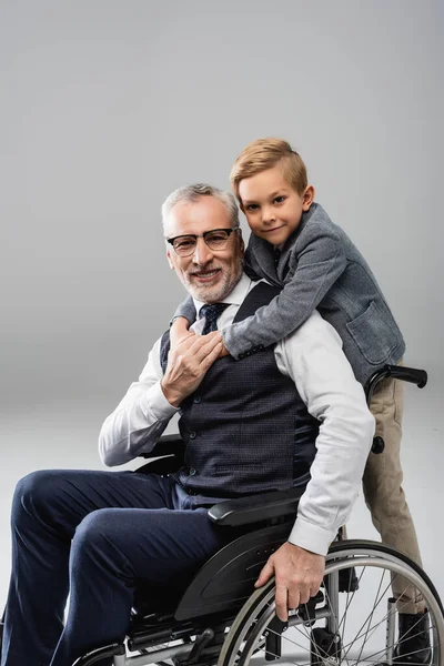 Lächelnder Junge umarmt Großvater im Rollstuhl, während er gemeinsam auf grau in die Kamera blickt — Stockfoto
