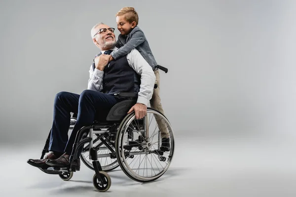 Happy man in wheelchair looking at smiling grandson hugging him on grey — Stock Photo