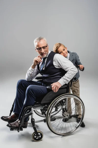 Smiling boy leaning on back of grandfather in wheelchair on grey — Stock Photo