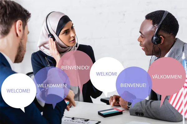 Interracial business partners in headsets near digital translators and interpreter, speech bubbles with word welcome in different languages illustration — Stock Photo