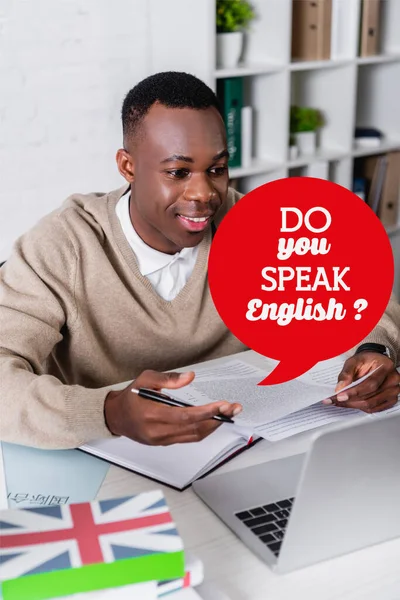 Feliz intérprete afroamericano trabajando cerca de la computadora portátil en primer plano borroso, burbuja de habla con habla Inglés pregunta ilustración - foto de stock