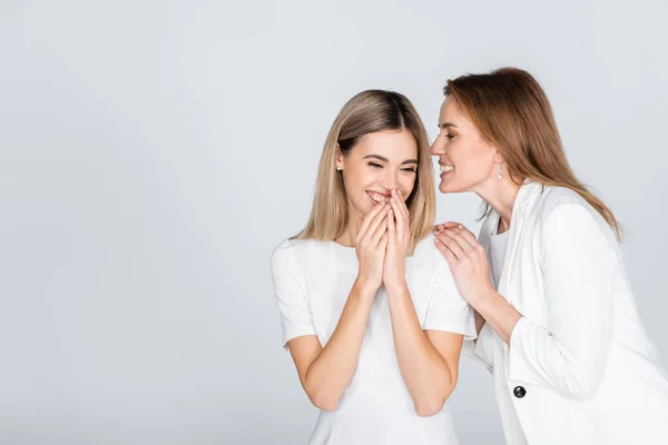 Cheerful mother whispering in ear of smiling daughter isolated on grey — Stock Photo