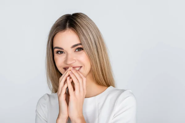 Cheerful young woman in white outfit smiling and covering mouth isolated on grey — Stock Photo