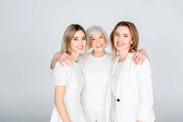 Три поколения веселых женщин улыбаются, глядя в камеру, и обнимаются на сером фоне — стоковое фото