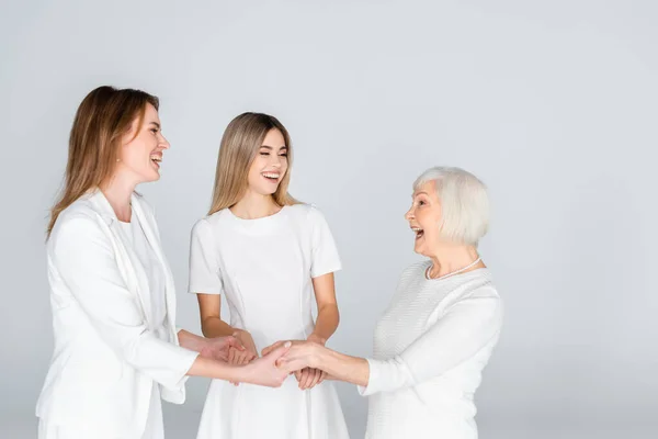 Tres generaciones de mujeres alegres sonriendo mientras se toman de las manos aisladas en gris - foto de stock