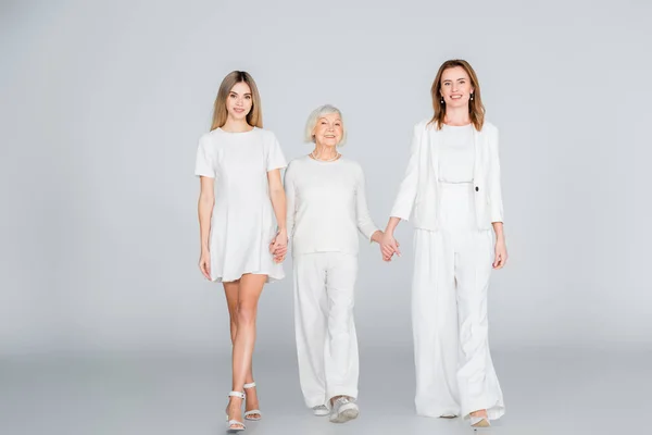 Повна довжина трьох поколінь щасливих жінок, які посміхаються, тримаючись за руки і ходячи на сірому — стокове фото