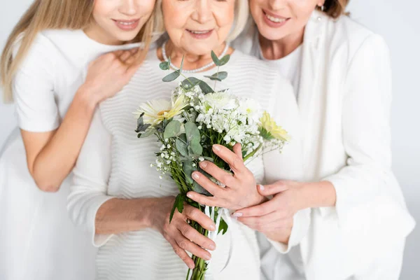 Vista recortada de la mujer mayor complacida sonriendo mientras sostiene flores cerca de hija y nieta aislada en blanco — Stock Photo