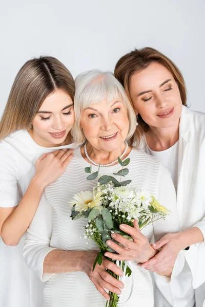 Felice donna anziana sorridente mentre tiene fiori vicino figlia e nipote isolato su bianco — Foto stock