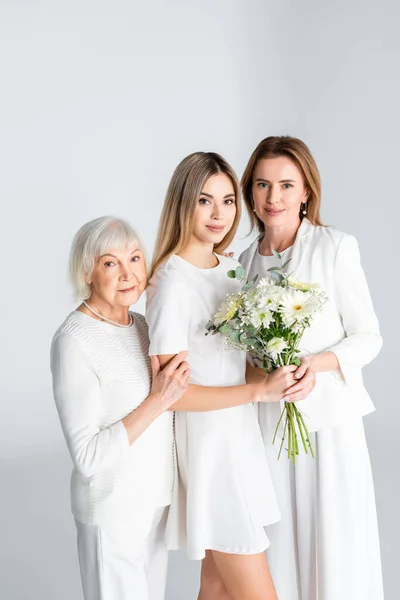Happy young woman smiling while holding flowers near mother and granny isolated on grey — Stock Photo