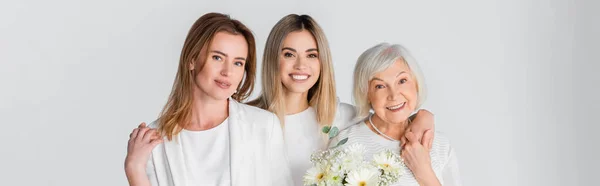 Drei Generationen glücklicher Frauen lächeln, während sie sich in der Nähe von Blumen umarmen, isoliert auf grauem Banner — Stockfoto