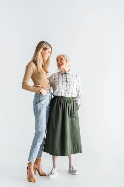 Longitud completa de alegre nieta y abuelita de edad avanzada de pie con las manos en los bolsillos en blanco - foto de stock