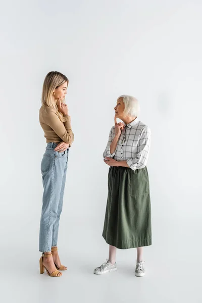 Longitud completa de la nieta reflexiva y abuelita mayor de pie y mirándose el uno al otro en blanco - foto de stock