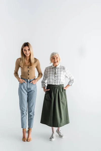 Повна довжина щасливої онуки і бабусі, що стоїть з руками в кишенях на білому — стокове фото
