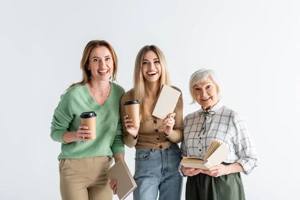 Tres generaciones de mujeres alegres sosteniendo vasos de papel y libros aislados en blanco - foto de stock