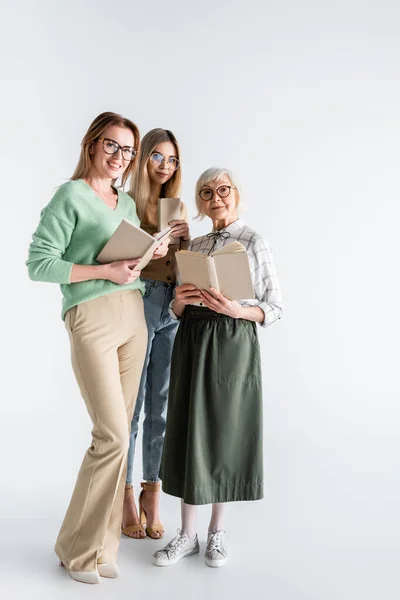 Longitud completa de tres generaciones de mujeres sonrientes en gafas con libros en blanco - foto de stock